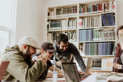 Grup de studenți care colaborează zâmbind în jurul unui laptop, folosind instrumente AI pentru proiectele universitare.