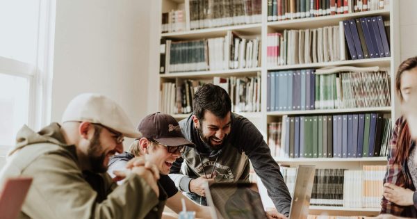 Grup de studenți care colaborează zâmbind în jurul unui laptop, folosind instrumente AI pentru proiectele universitare.
