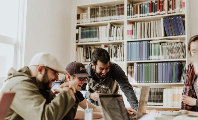 Grup de studenți care colaborează zâmbind în jurul unui laptop, folosind instrumente AI pentru proiectele universitare.